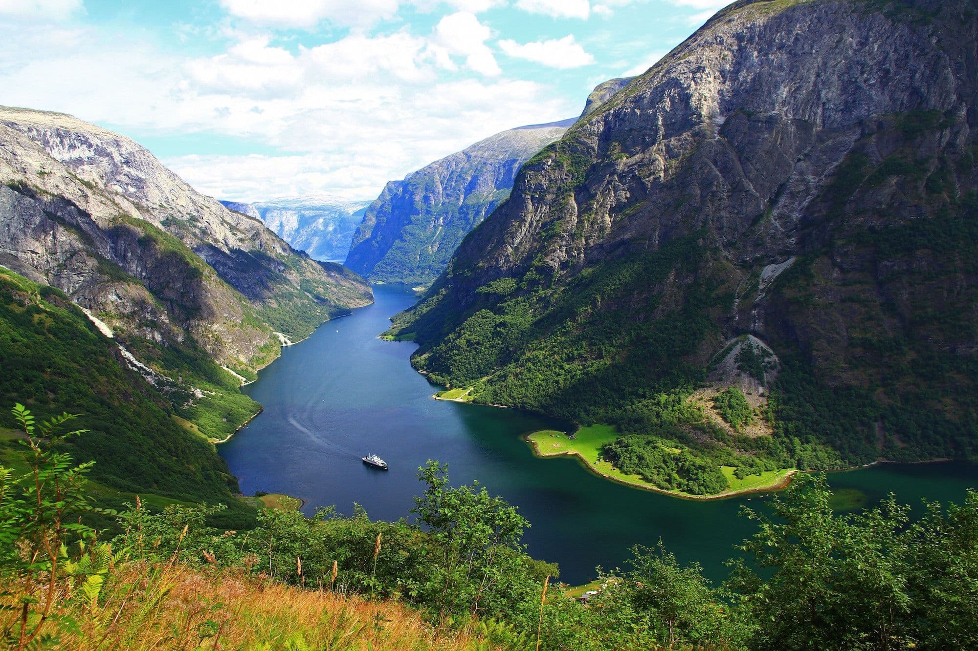 Panoramic view of blue fjord winding through rocky mountains, lush greenery, small cruise ship sailing, and green grassland.
