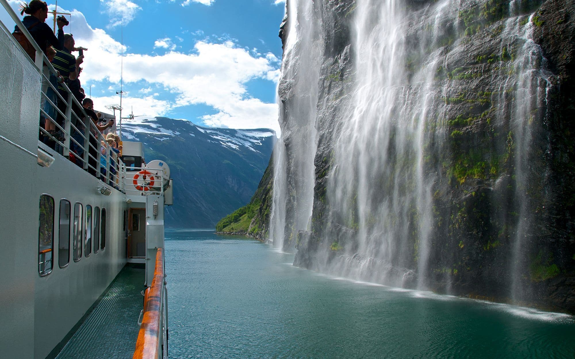 Kryssingskip seiler gjennom fjord med foss som renner fra høy fjellvegg, reflekterende vann og en rolig atmosfære.