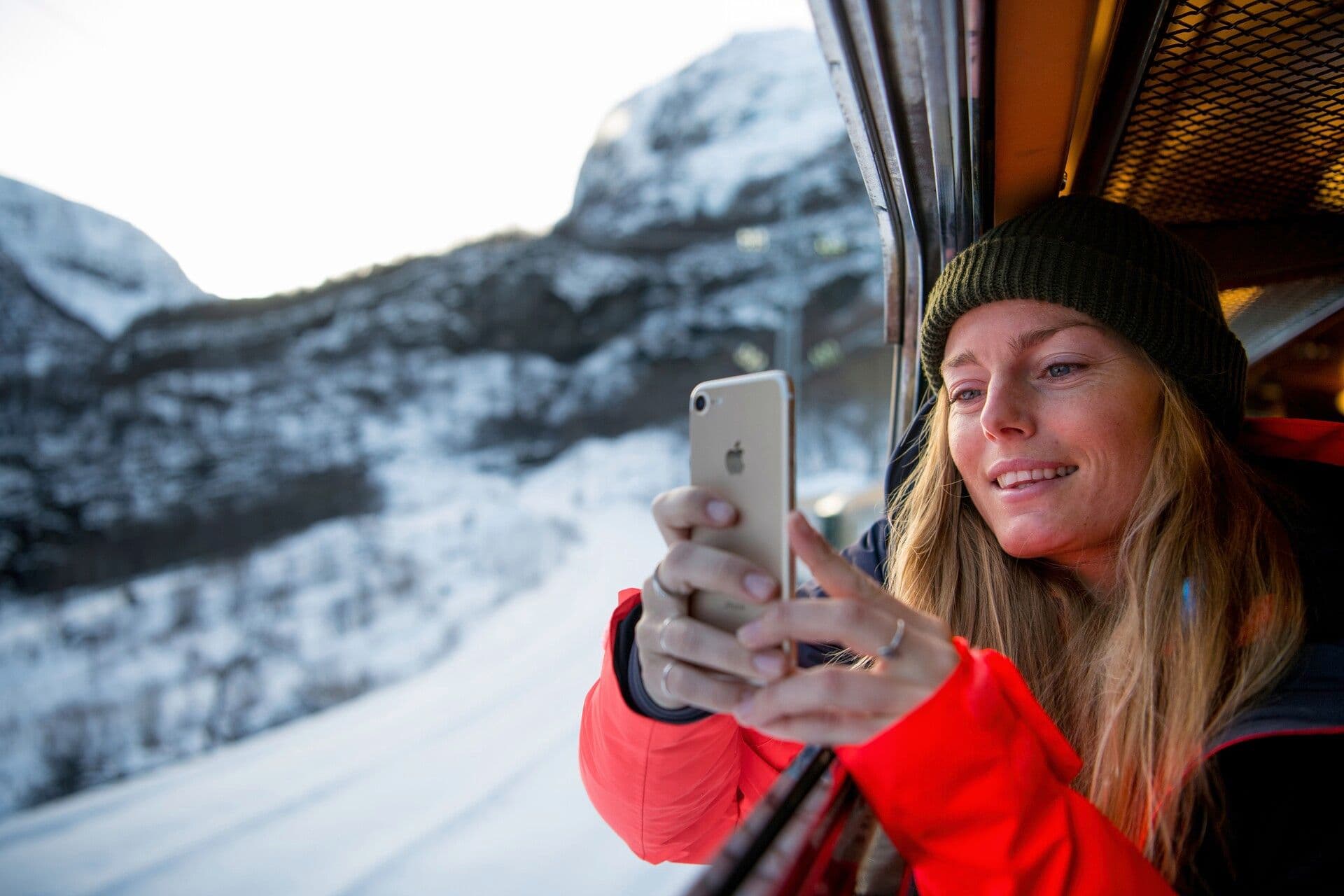 Woman in red jacket and green beanie takes photo from train window.