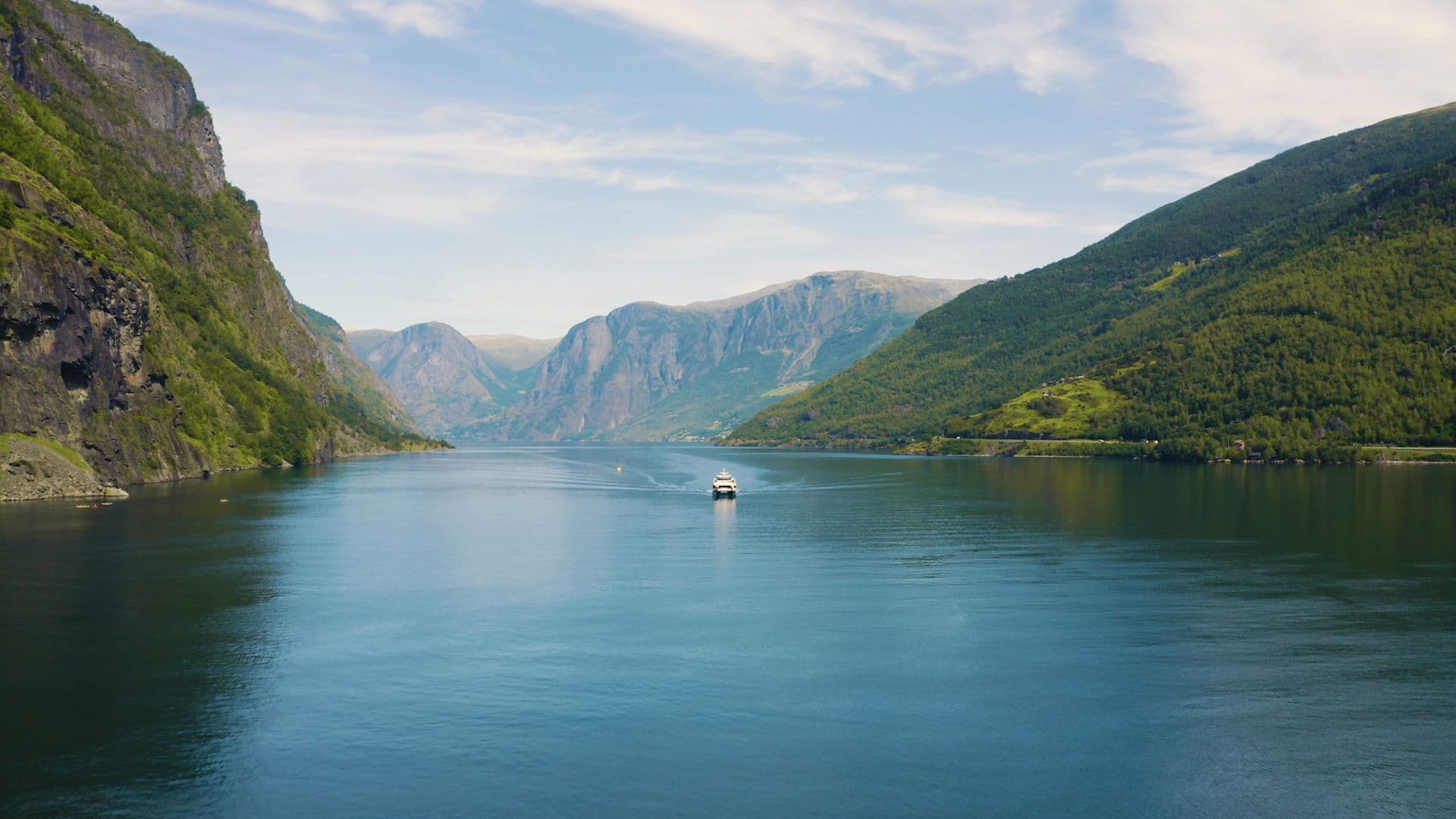 Boat cruises through calm Sognefjord surrounded by green cliffs.