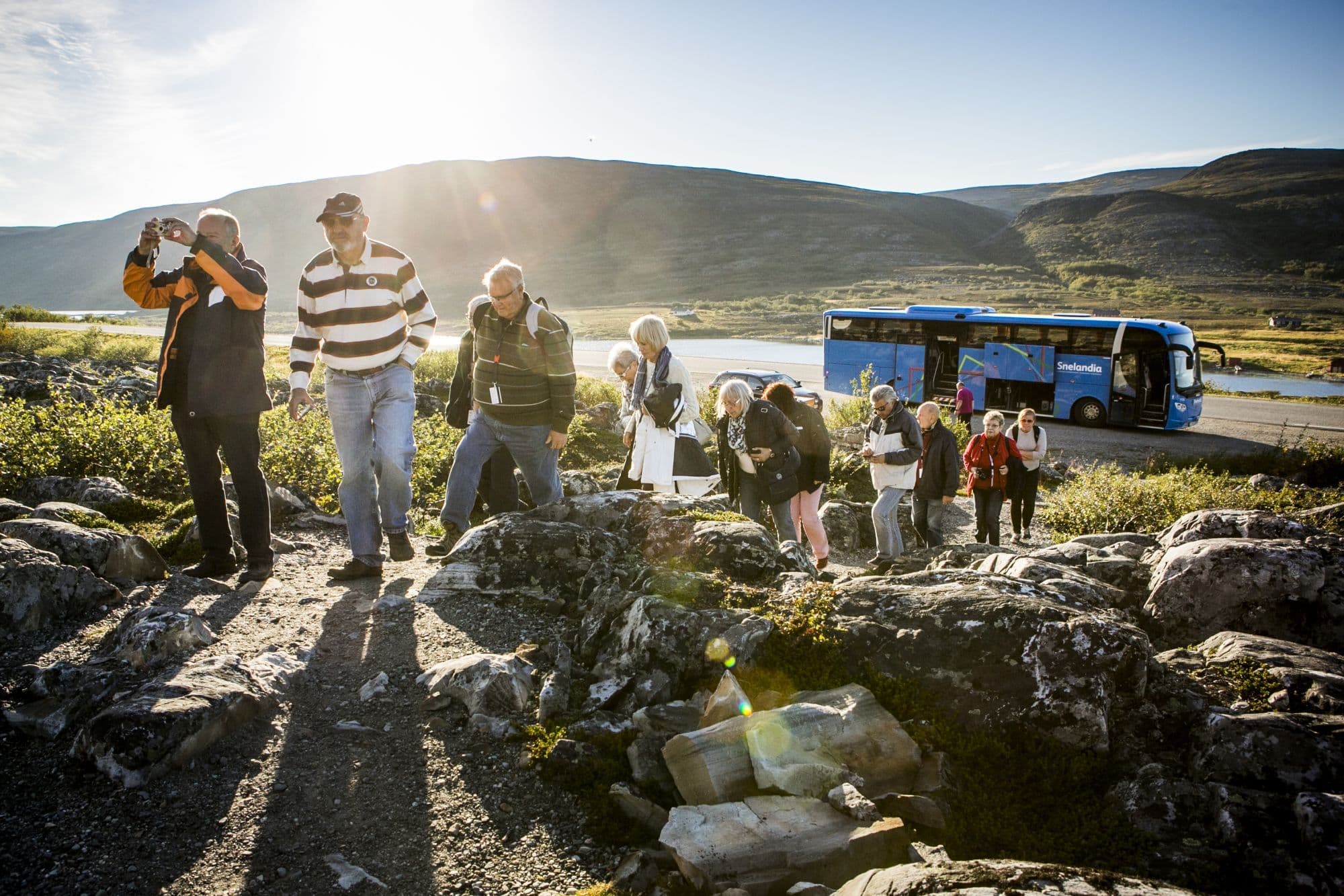 Turister går av blå buss for fottur i steinete fjell i gyllent ettermiddagslys.